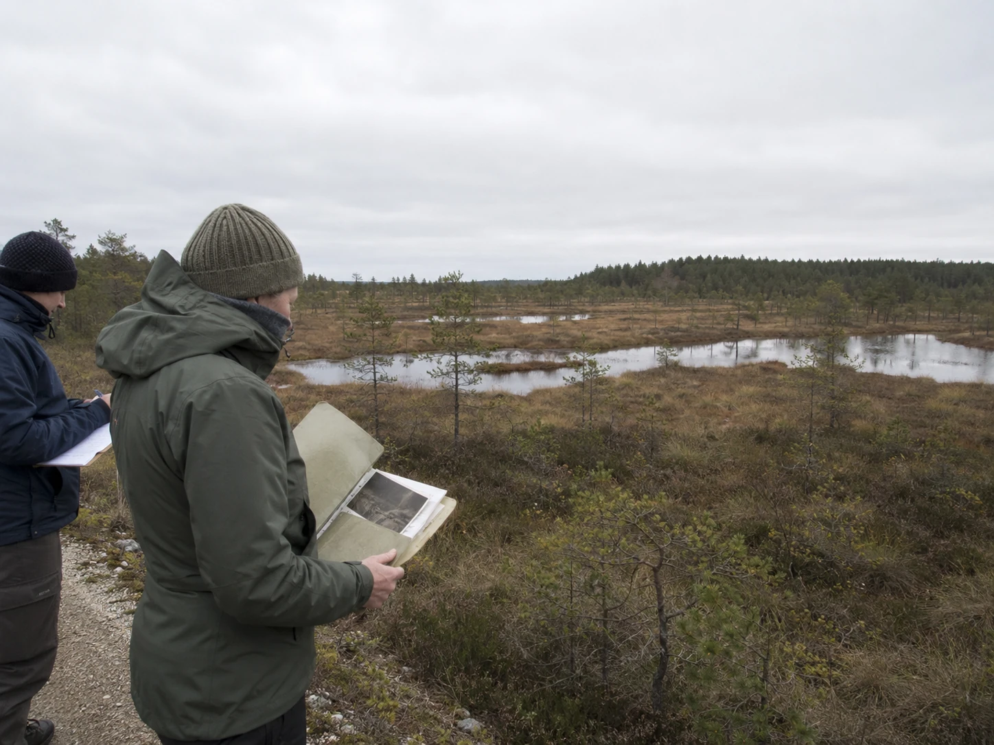 Two practitioners in the field compare a historic photograph from an album against the present wetland landscape while recording notes.